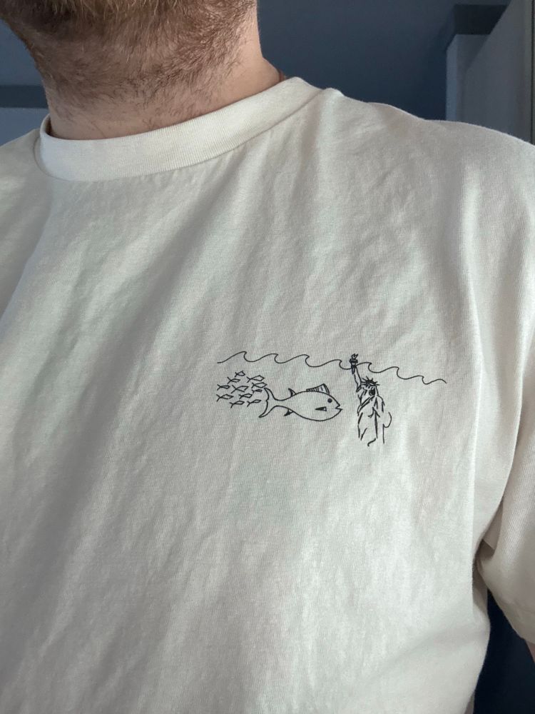 A shirt with a picture of the Statue of Liberty sunken into the ocean. Fish swim around her. She is dead