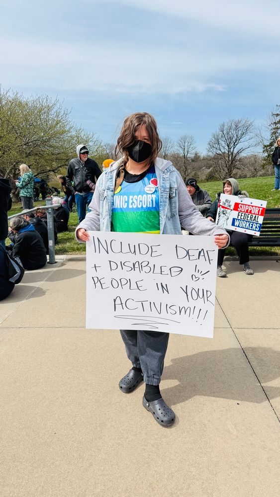 Rikki at protest with sign that says "include deaf and disabled people in your activism!”