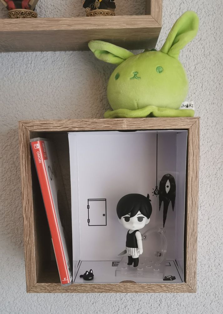 La Nendoroid de Omori dans son décor de la White Room placé sur étagère mural à part. Le jeu physique et une petit peluche de slime bunny vert sont rangés avec. 