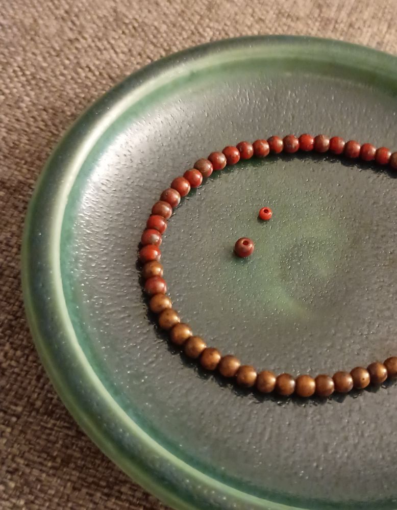 錆色と赤＋緑の2色ビーズ（3mm）のブレスレットを濃緑の小皿の上に置いてあります