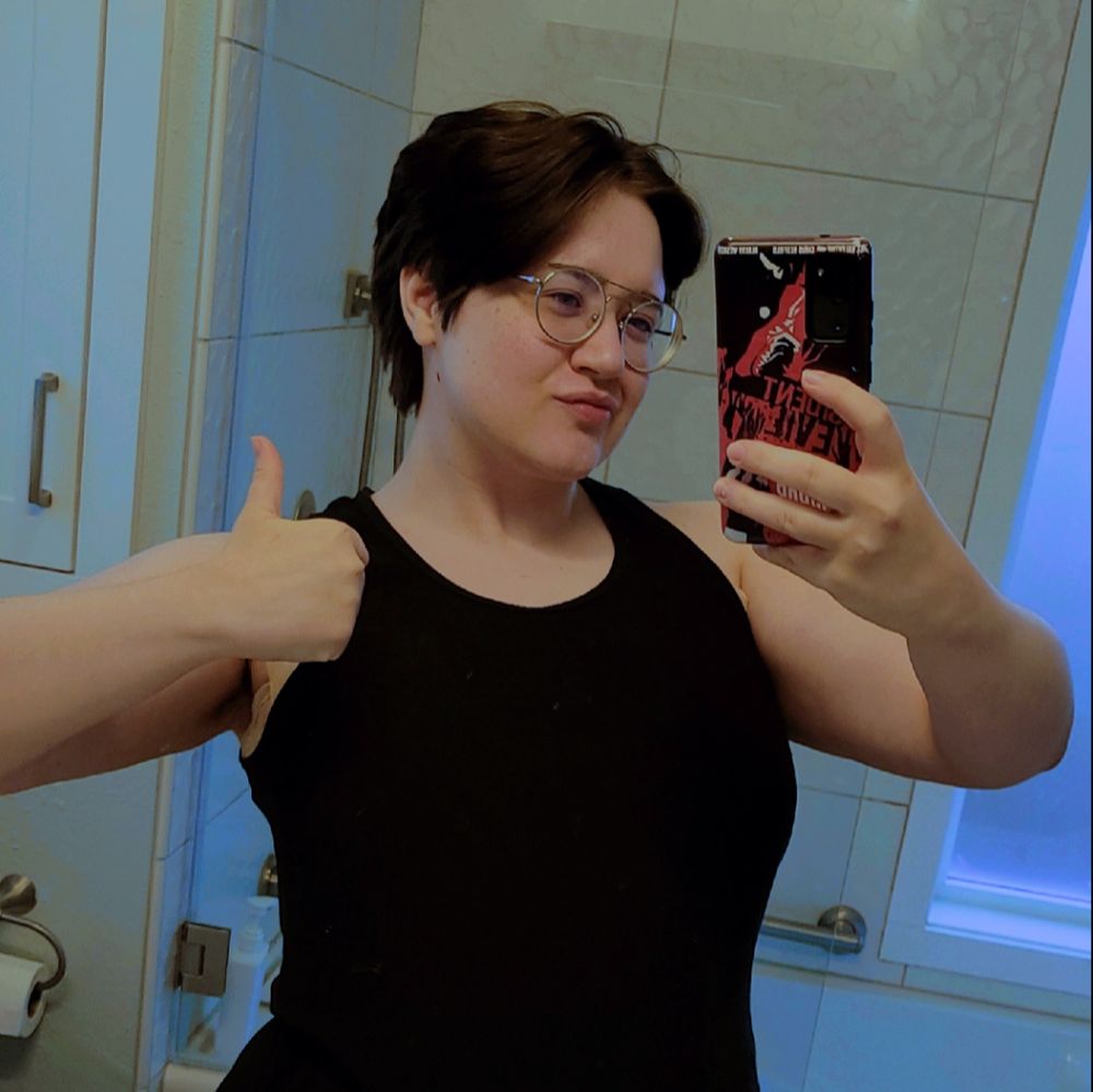 A bathroom mirror selfie of Angstmilk giving a thumbs up, wearing a blank tank top. 