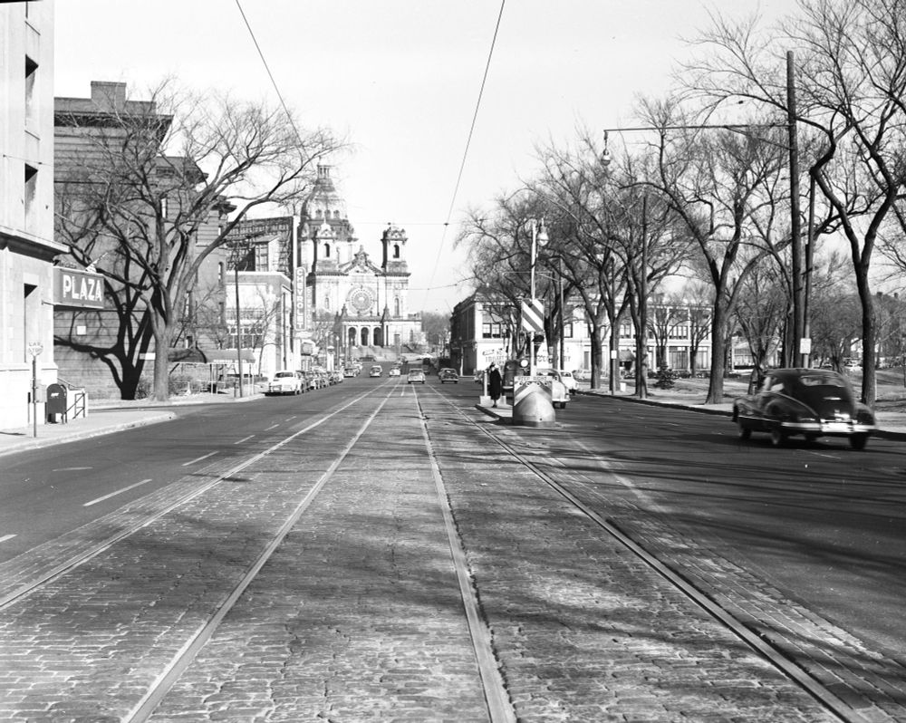 Black and white photo of Hennepin Avenue early spring, with trees on either side. Loring Park is on the left, a variety of brick and stone apartment and commercial buildings on the right. The facade of St Mary's Cathedral is in the distance in center view. Streetcar tracks and a streetcar stop in middle. 1952
