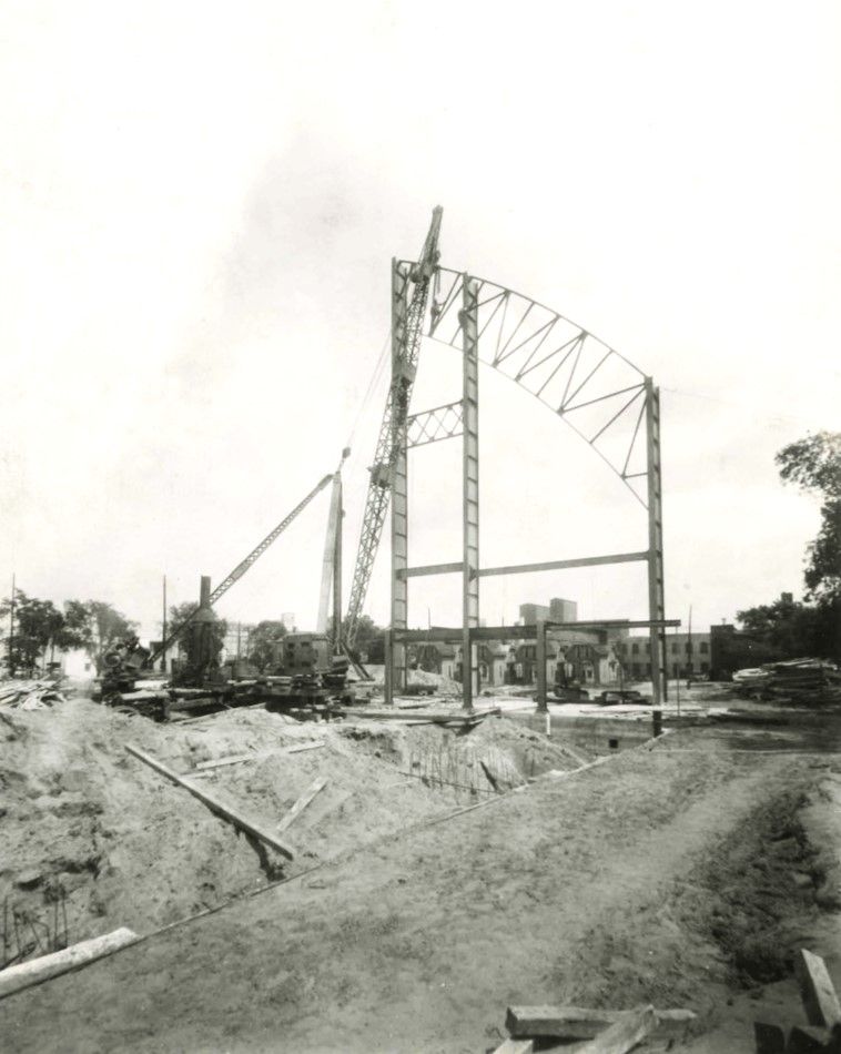 3 single columns with a 1/2 piece of arched steel rise high above dirt piles with the assistance of a steam crane. Pile drivers can be seen on the left. University of MInnesota LIbraries photo. 