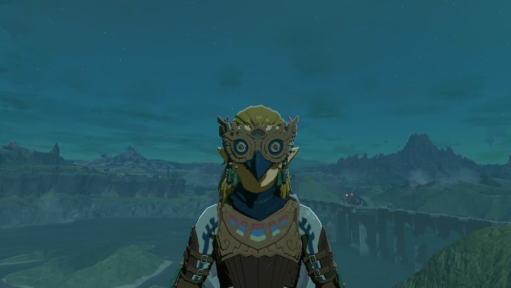 Link from Tears of the Kingdom, dressed in the Glide Suit; notably, the mask has very large bird-like eyes. He is staring directly at the viewer. In the background is the Bridge of Hylia Flame Gleeok.