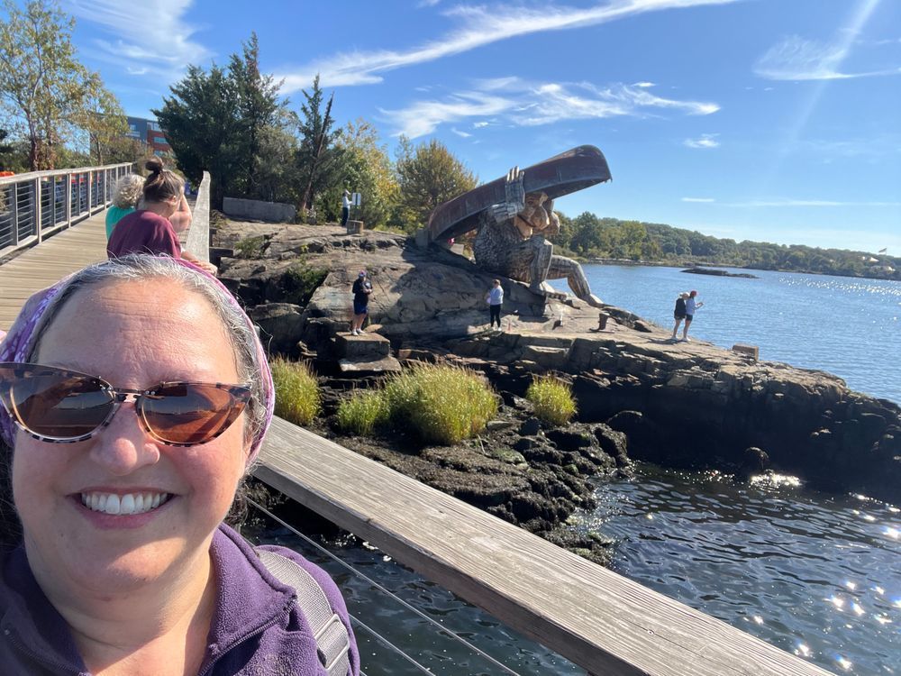 Rep. Kislak selfie in sunglasses in front of large wooden troll holding a boat over the head on a rock next to the water with people standing near it.
