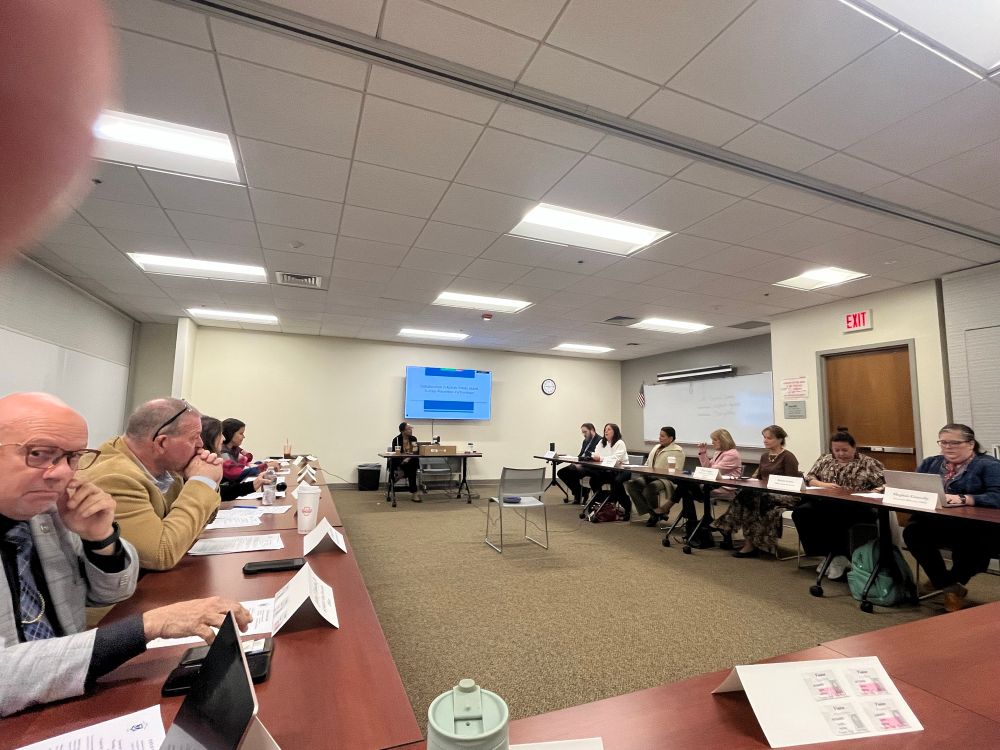 Picture of ppl in a room around a horseshoe shaped table. 