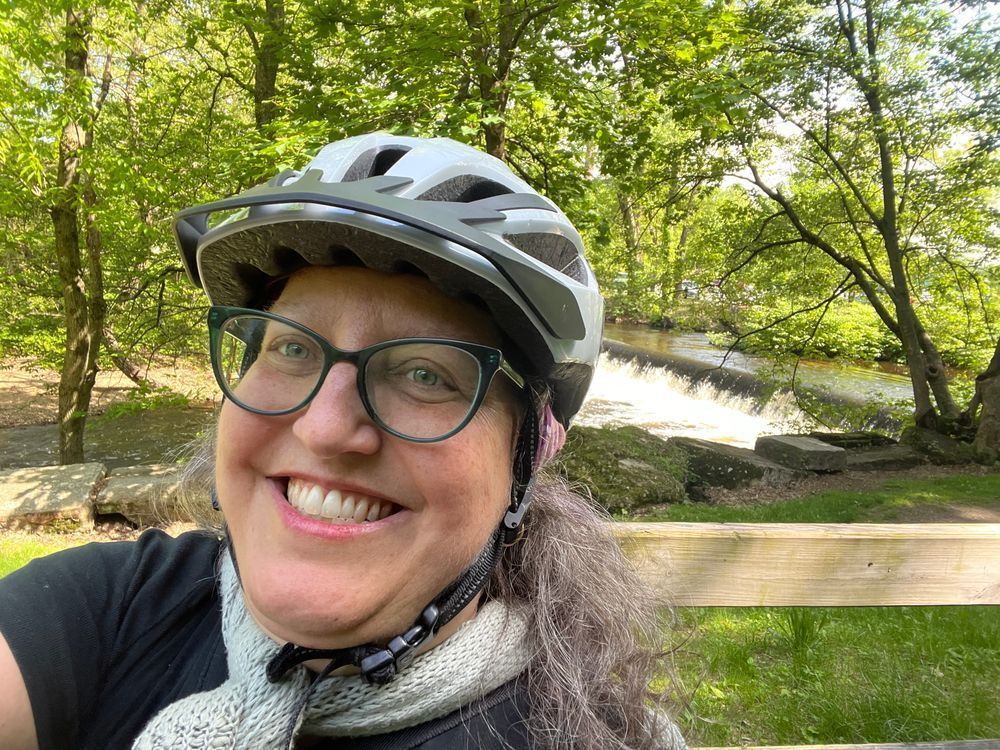 smiling woman with glasses and bike helmet, with little waterfall behind