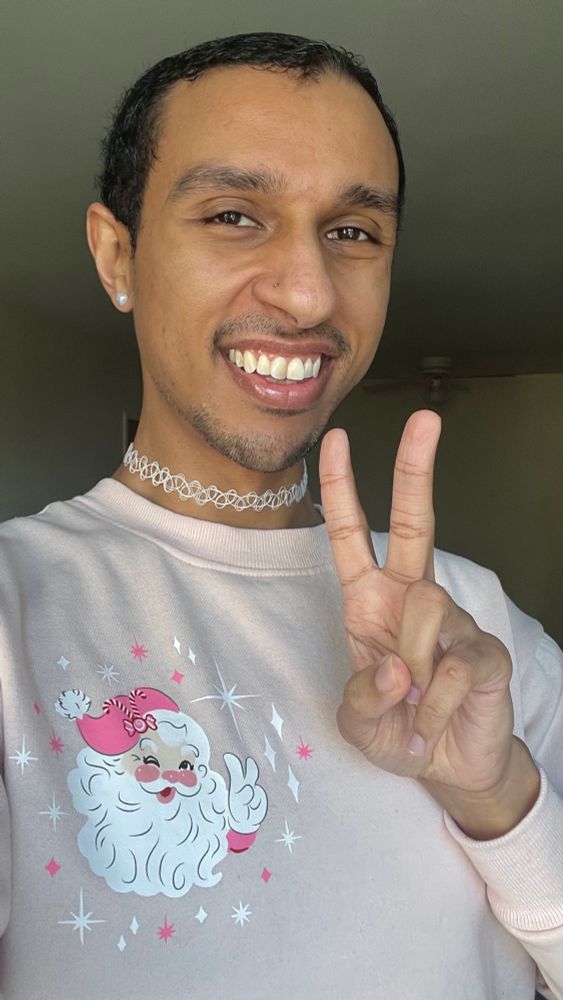 A photo of me smiling & making a peace sign while wearing a white choker & pink sweatshirt with a Santa Claus face making a peace sign & smiling.