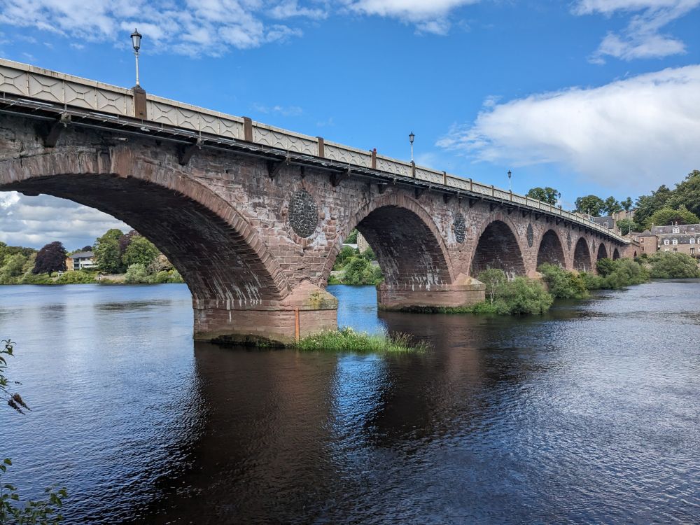 A stone bridge with more than five arches crosses a wide river under a blue sky.
