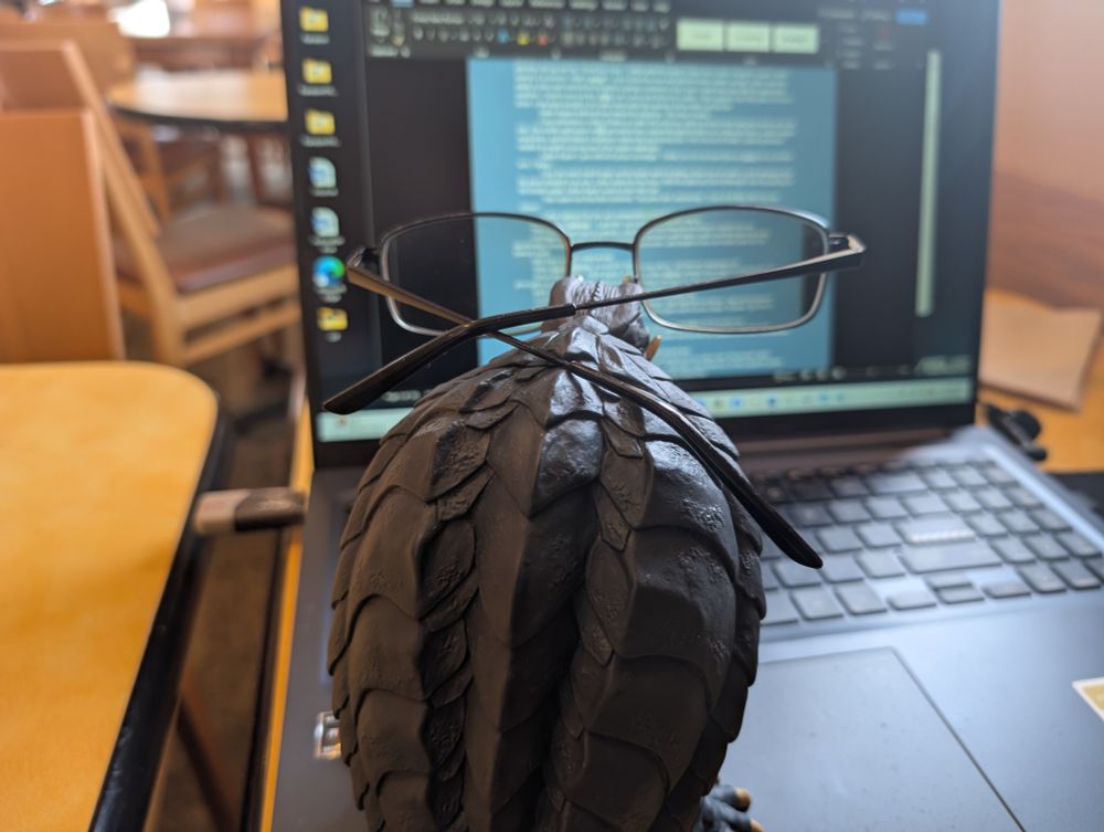 A toy Gamera (A giant fire-spewing turtle kaiju) standing on an open laptop wearing regular human glasses appearing to read an open document on the screen.
