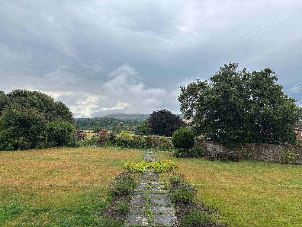 A complex of contrasting grey and white clouds rolling over Firle Beacon. A parched lawn damp from one night of rain. Green trees and bushes. A flagstone path with a central sundial, lavender to each side. 