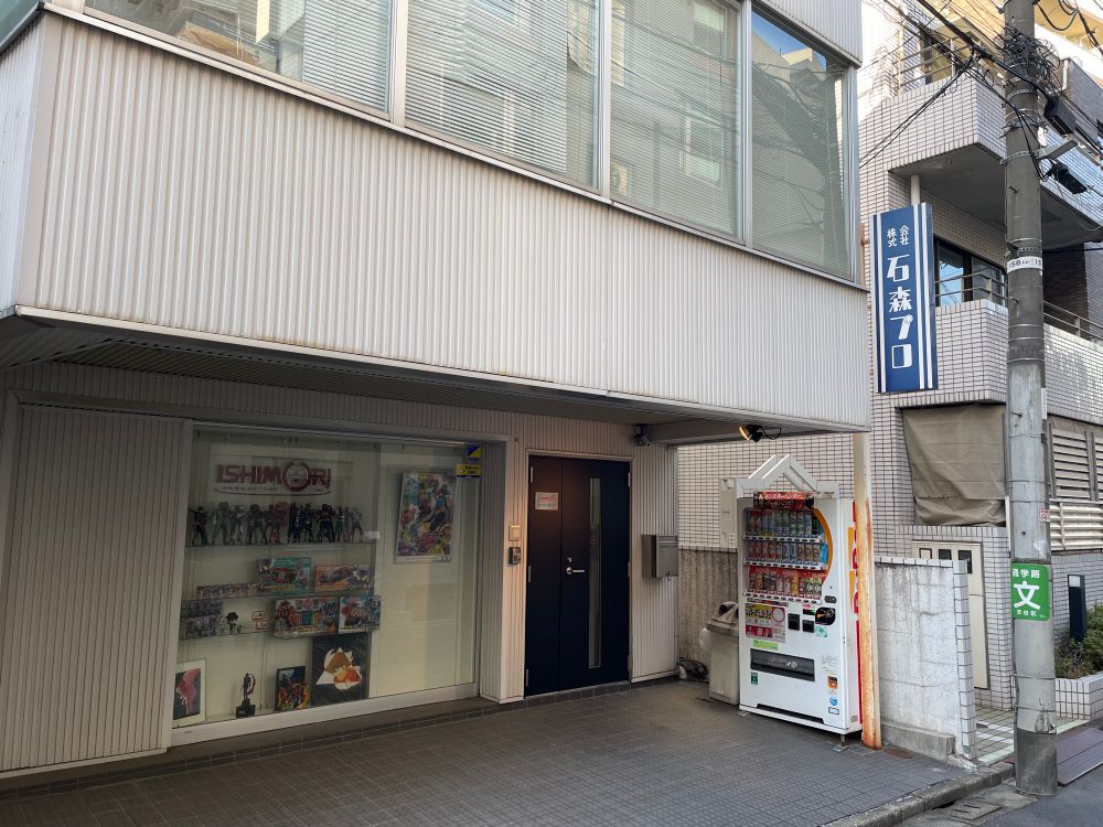 Front of an office building. On the right is a Japanese sign saying Ishimori Pro. Display shelves in the window show Kamen Rider and Cyborg 009 merchandise. Between that and the front door is a poster for the current Kamen Rider series, Gabu / “Gulp”.