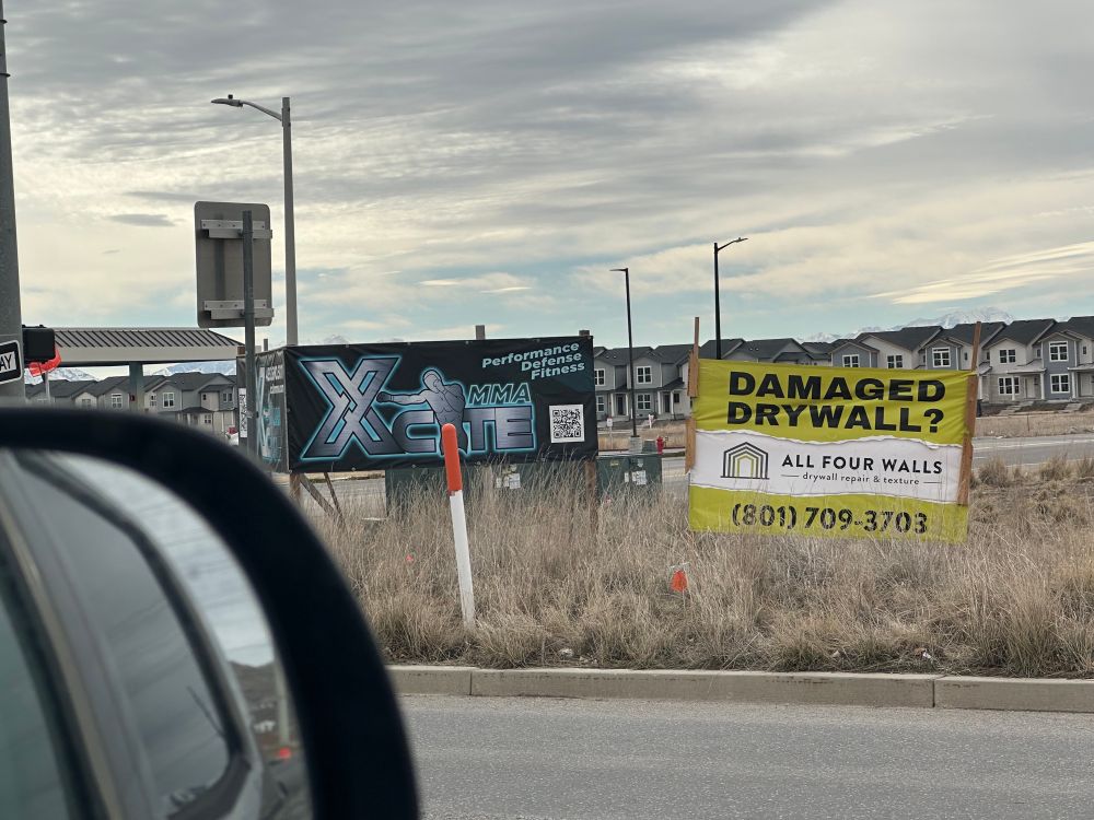 two signs next to each other. the one on the left is for a company called “MMA Xcite” offering self defense training, the one on the rights says “damaged drywall?” advertising for a drywall repair company named all four walls