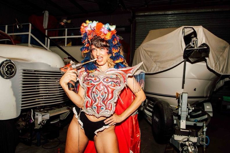 A colourfully dressed performer stands in front of a boat and an old car with a drill in their mouth while wearing a large head piece and a red cape.