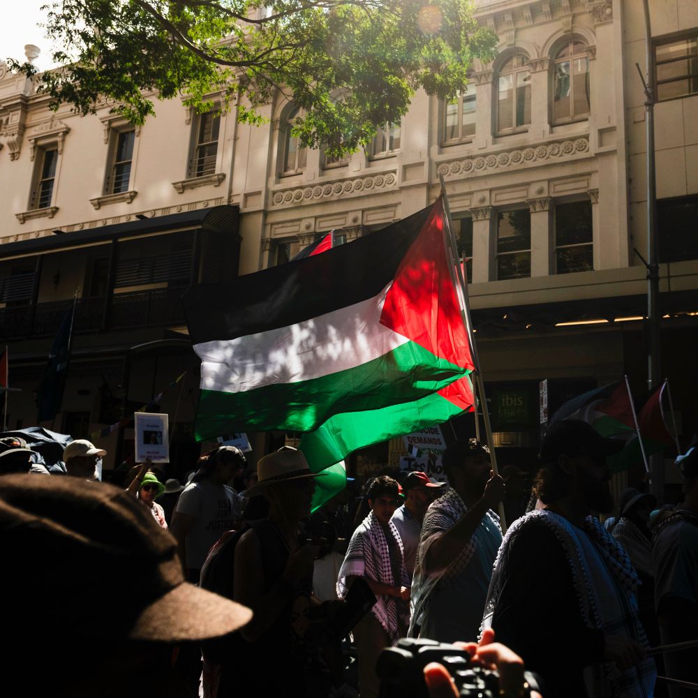 A photograph of a pro Palestinian march on the streets of so called Brisbane. The photo features protesters holding a group of Palestinian flags