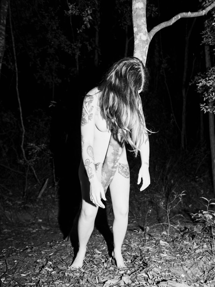 Black and white flash photograph of a nude femme model draped on the trunk of a skinny tree. 