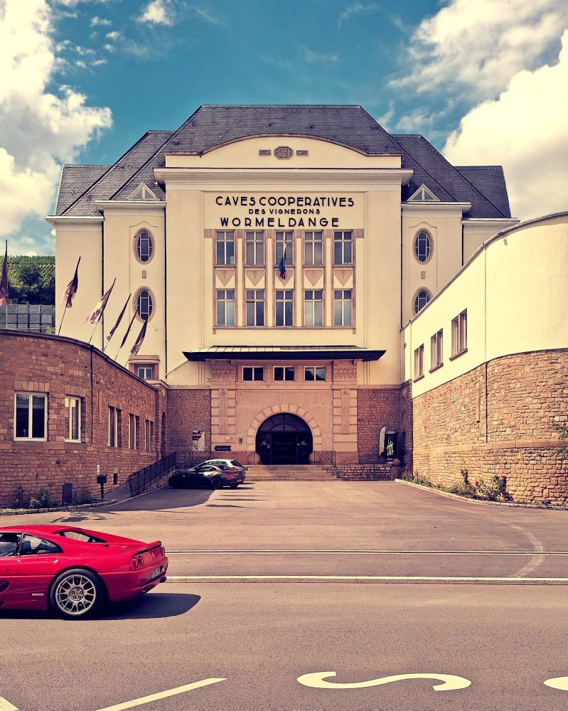 Frontansicht auf das 1931 am Moselüfer fertig gestellt Weinkellergebäude. Der Sockel ist Sandstein ausgeführt und wirkt mit dem Eingangsportal wie eine asiatische  Stadtmauer. Im Vordergrund ist das rote Heck eines Ferrari F355 zu sehen.