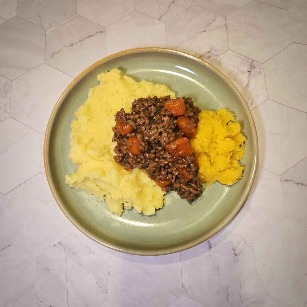 A green plate on a marble backdrop. The plate has on it some mashed potatoes and mashed Swede topped with some minced beef and carrots. The photo is shot from above