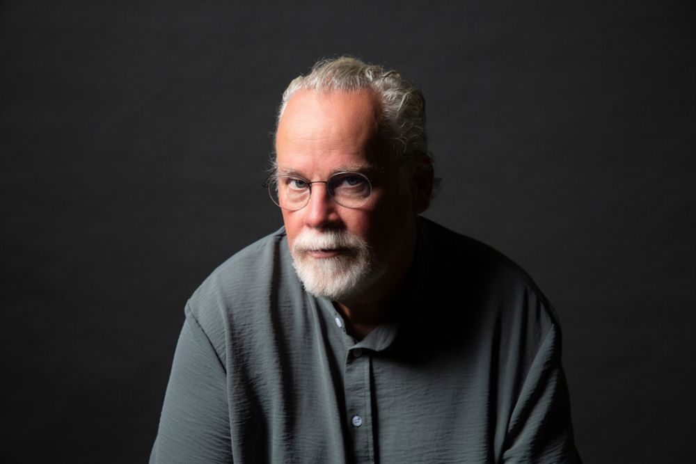 This is a photo of international bestselling author Michael Connelly. He’s sat starring at the camera, wearing a loose grey shirt, looking serious - but cool. 

He wears glasses, has a white bushy goatie beard and grey hair slicked back. 