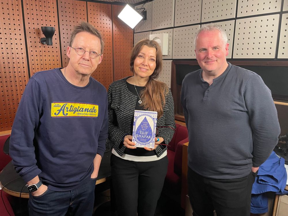Simon Mayo is on the left of the image, Elif Shafak is in the middle and Matt Williams is on the right. They are standing in a recording studio looking at the camera. Elif holds her new book “There Are Rivers In The Sky”