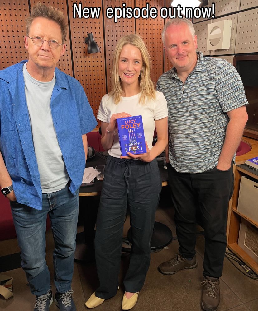 The photo is of three people standing in a podcast studio.

On the left of the image is broadcaster Simon Mayo, who has his hands in his pockets and is looking directly at the camera with a slight smile. He’s got a blue shirt on and jeans.

In the middle is the author Lucy Foley. She’s holding up a copy of her new book ‘The Midnight Feast’ and is looking directly at the camera too. She’s wearing black trousers and a white top. 

On the right hand side is broadcaster Matt Williams. He’s smiling at the camera, with his hands in his pockets, and is wearing a grey top and black jeans.