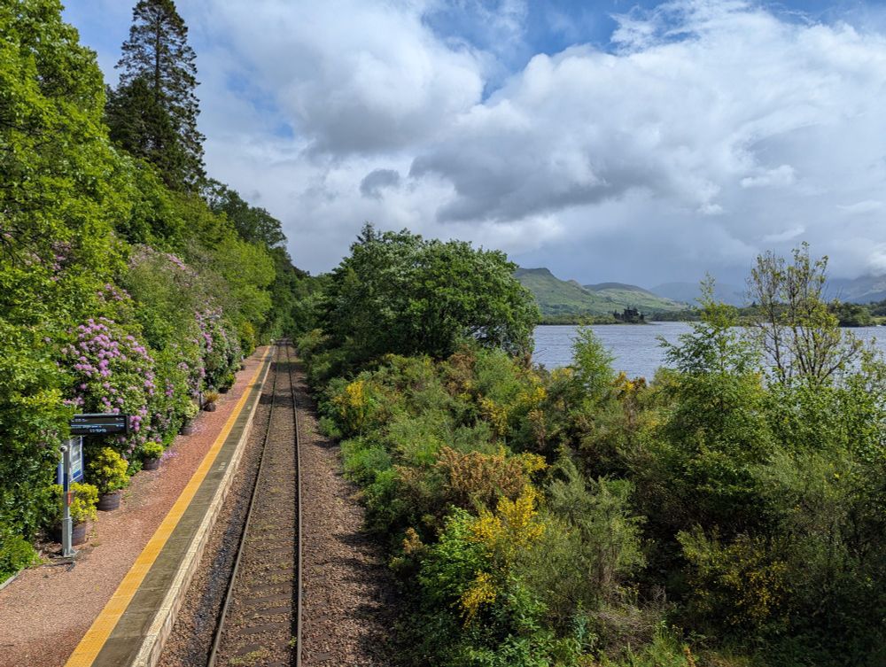 View from the footbridge, single line and platform to the left, loch to the right with a castle in the distance 
