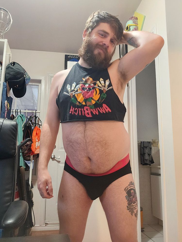 Rexxx is standing facing the camera in a tight, black cropped tank top with Scary Terry from Rick and Morty on it. He's also wearing a black brief with a hot pink waistband.