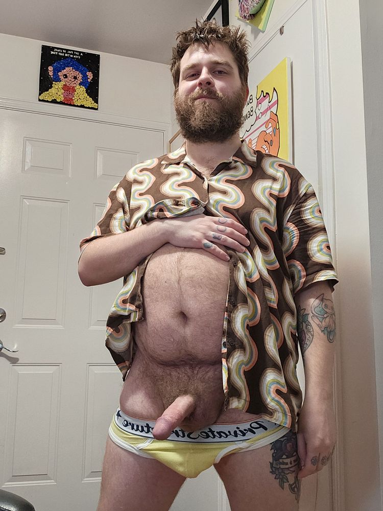 Rexx is standing facing the camera. He's wearing a yellow vintage brief with white trim, and a brown, tan, pink and teal patterned button up. The briefs are pulled down and his hard cock is out.
