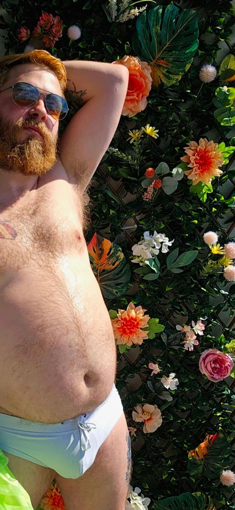 Rexxx is standing in front of a brick wall and a lattice covered in ivy and fake white, yellow , pink and coral flowers. He is wearing a sheer, stripped green beach robe, a white speedo and aviator sunglasses. His belly listens with sweat in the sunlight.