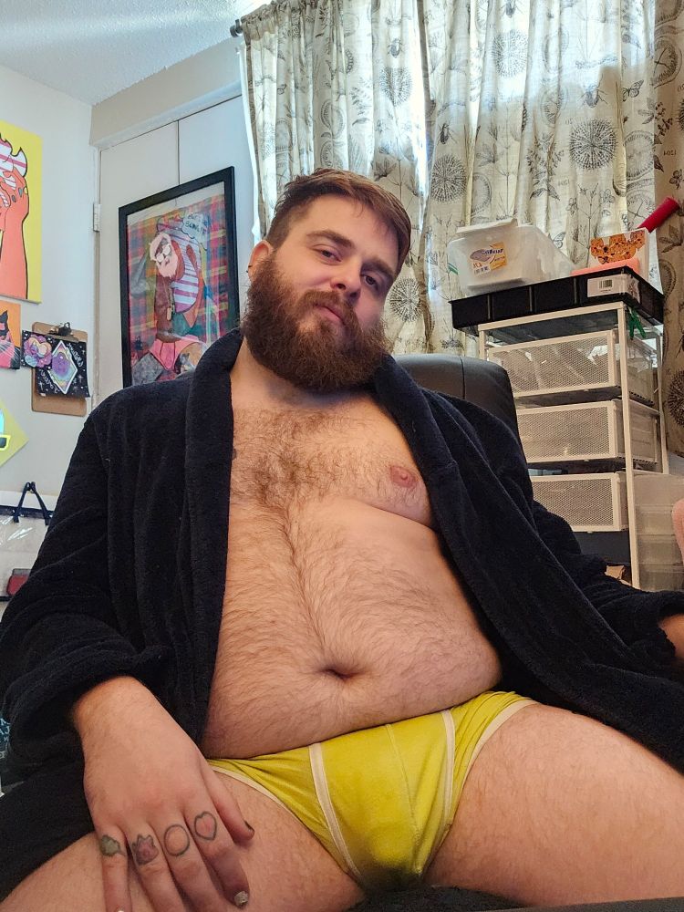 Rexxx is sitting in his office hair. His navy bathrobe flung open, partially exposing his chest. His neon yellow trucks with white trim are on full display, showing the outline of his cock.