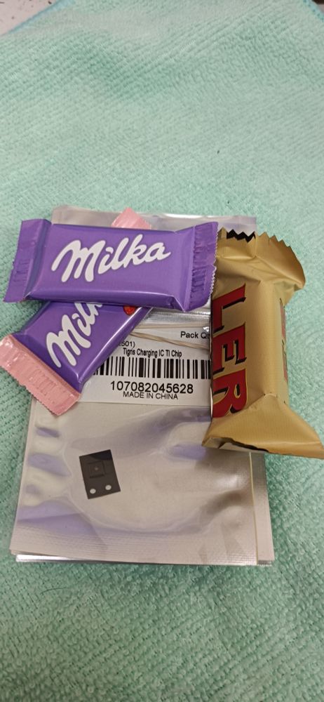Das Bild zeigt eine ungewöhnliche Kombination aus Elektronik und Süßigkeiten. Im Zentrum liegt eine metallisch glänzende Verpackung, wie sie typischerweise für empfindliche elektronische Bauteile verwendet wird – antistatisch und mit einem Barcode versehen. Auf dem Etikett ist unter anderem zu lesen:

„Charging IC Chip“ – ein Hinweis darauf, dass sich im Inneren ein Ladechip befindet, vermutlich für elektronische Geräte.

„MADE IN CHINA“ – Herkunftsangabe.

Eine lange Nummer: 1xxxxx – vermutlich eine Serien- oder Produktnummer.

In der transparenten Ecke der Verpackung ist ein kleiner schwarzer Chip sichtbar – das technische Herzstück.

Doch das eigentlich Skurrile: Obenauf liegen drei Süßigkeiten – zwei Milka-Schokoladen in lila Verpackung und ein Toffi in gold-rotem Papier. Die Süßigkeiten wirken wie beiläufig abgelegt, fast wie eine kleine Belohnung oder ein ironischer Kontrast zur nüchternen Technik.