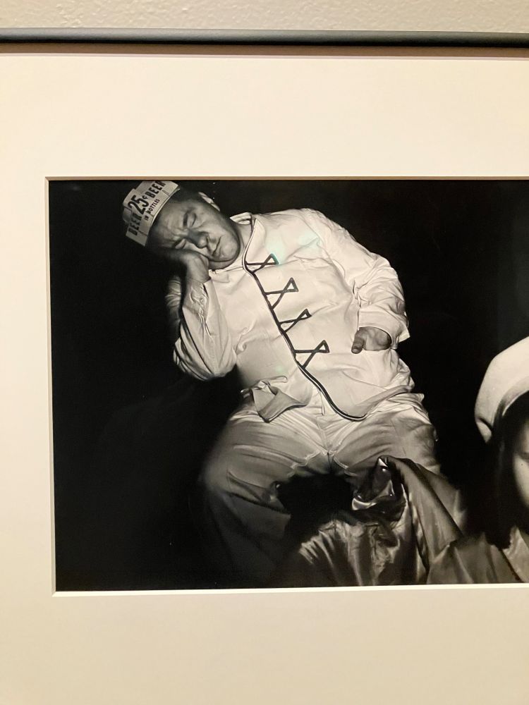 a black and white image of a white man in a white uniform with a hat that indicated he's selling beer for 25 cents. the man is asleep while sitting down and is leaning to one side, with his cheek resting against the palm of his hand
