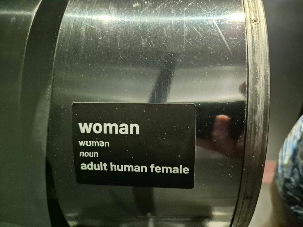 A toilet roll dispenser that has a sticker on it. The sticker is the 'Woman - adult human female' meme common amongst transphobes and idiots. 