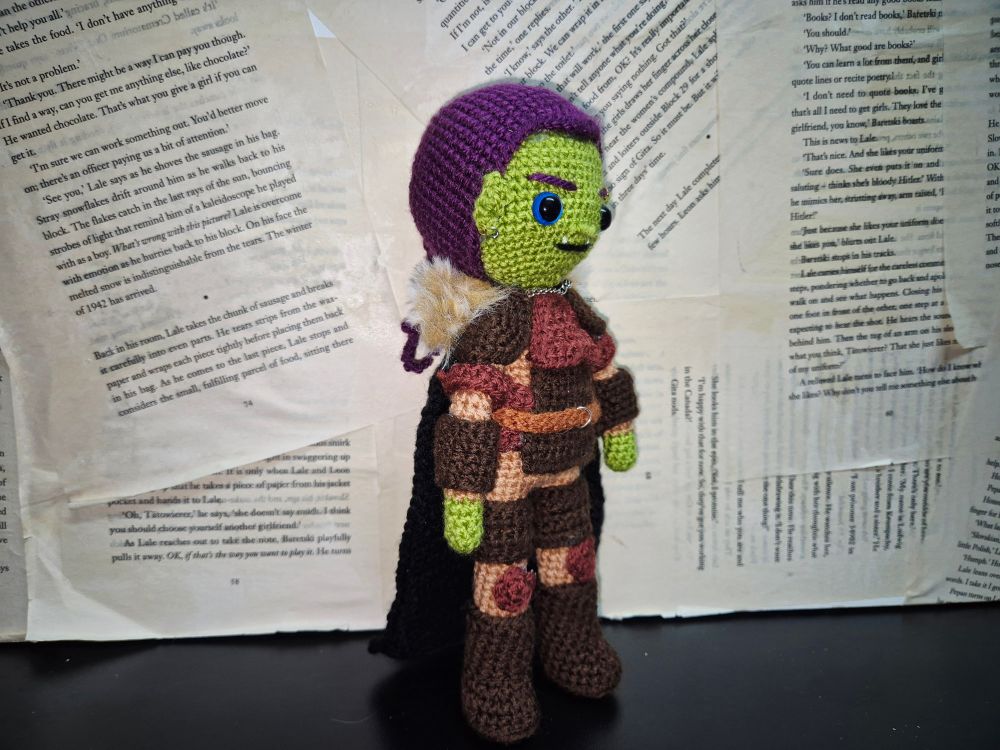 A hand made crochet doll of an original dungeons and dragons character. A green skinned half orc with purple hair tied back in a ponytail braid. She is wearing armour of various shades of brown, and a black cloak with a brown fur trim. She has some metal piercings on her ears and eyebrow. 