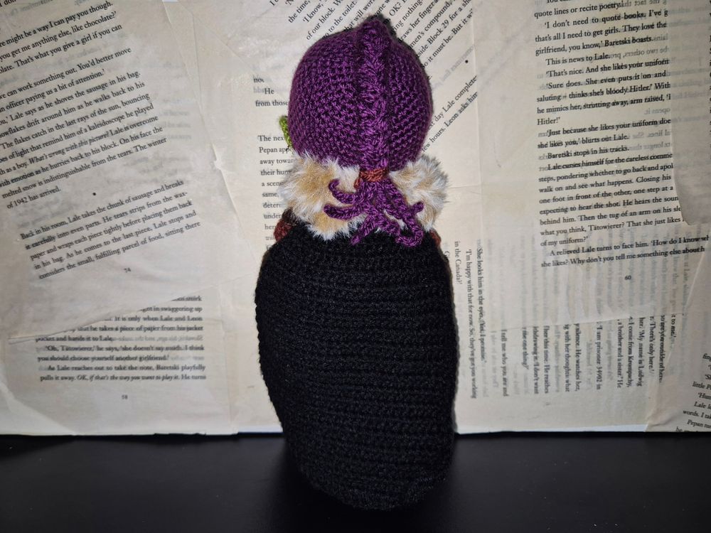 A hand made crochet doll of an original dungeons and dragons character. A green skinned half orc with purple hair tied back in a ponytail braid. She is wearing armour of various shades of brown, and a black cloak with a brown fur trim. She has some metal piercings on her ears and eyebrow. 
