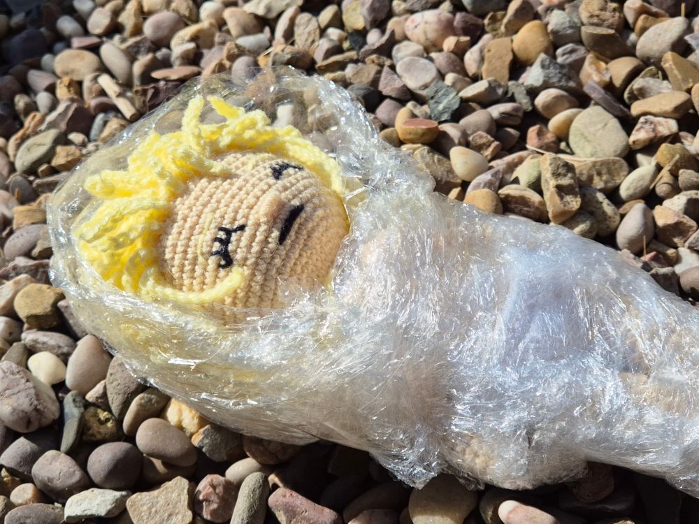 A crochet doll of the character Laura Palmer from the TV show Twin Peaks, as she is found dead in the first episode.

The dolls wrapped in plastic, with her face and hair uncovered. She has pale skin, blonde hair, and her eyes are closed. The doll is laying on gravel, depicting the scene in the first episode where she's found on a pebble lake shore. 