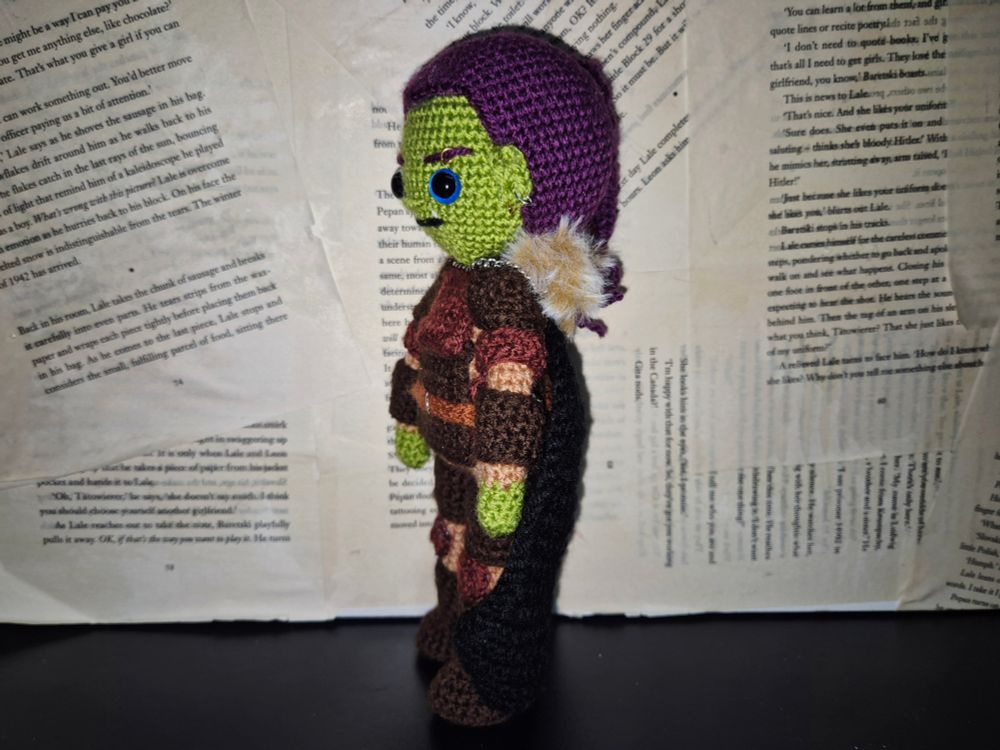 A hand made crochet doll of an original dungeons and dragons character. A green skinned half orc with purple hair tied back in a ponytail braid. She is wearing armour of various shades of brown, and a black cloak with a brown fur trim. She has some metal piercings on her ears and eyebrow. 