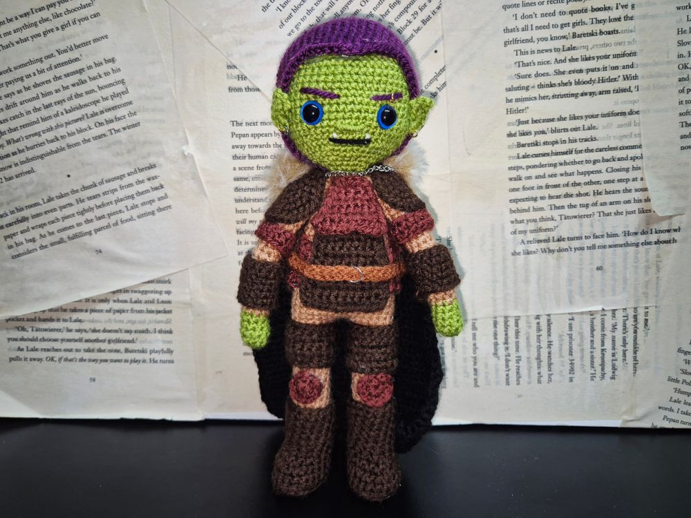 A hand made crochet doll of an original dungeons and dragons character. A green skinned half orc with purple hair tied back in a ponytail braid. She is wearing armour of various shades of brown, and a black cloak with a brown fur trim. She has some metal piercings on her ears and eyebrow. 