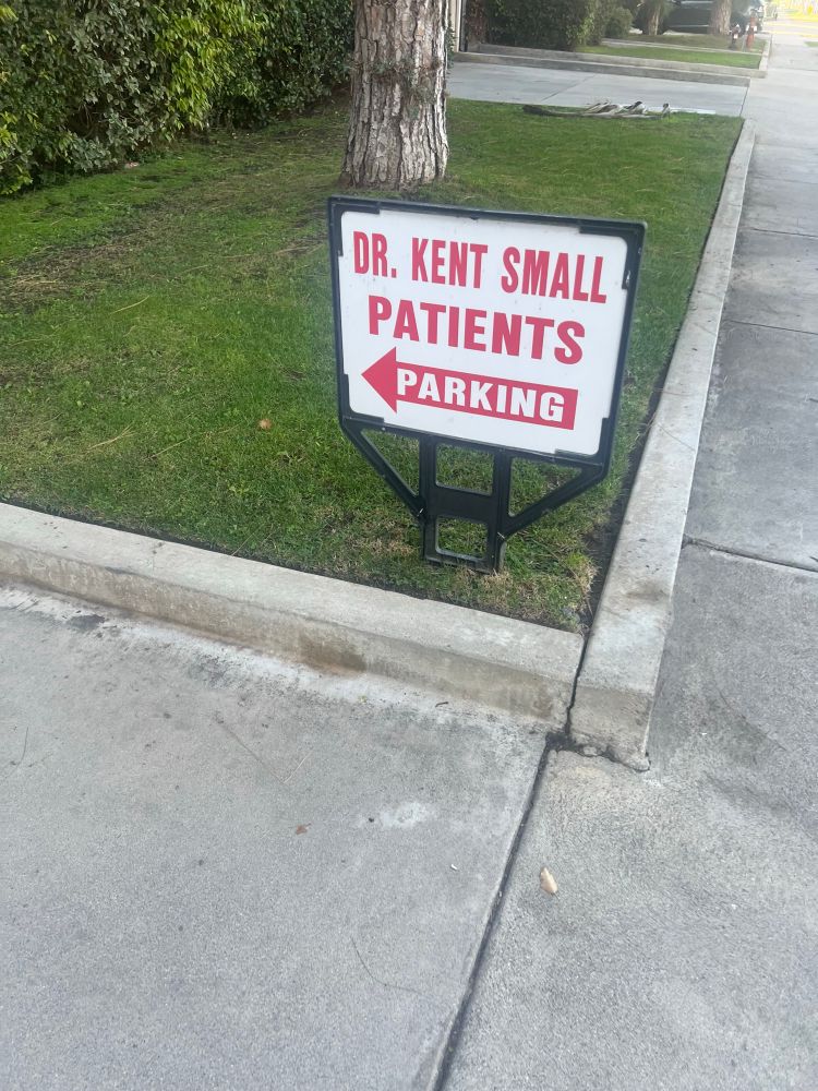 White sign with red letters, planted in the grass next to a driveway. The sign reads “Dr. Kent Small Patients Parking,” with a red arrow pointing to the left.”