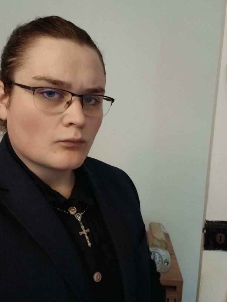 Ella is dressed formally in sermon attire for her weekly 'tea and cake with the vicar' community outreach programme. Black vicar garb with steel buttons, a steel crucifix hanging between.