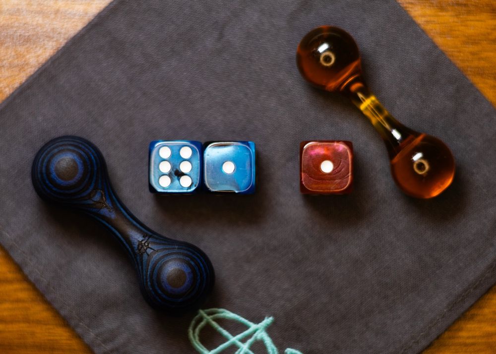 Three dice in the center of a grey handkerchief, with an amber ultem knuckle-roller on the top right and a black-and-blue spectraply knuckle-roller on the bottom left. The left two dice are blue and total 7, and the die on the right is amber and shows 1.