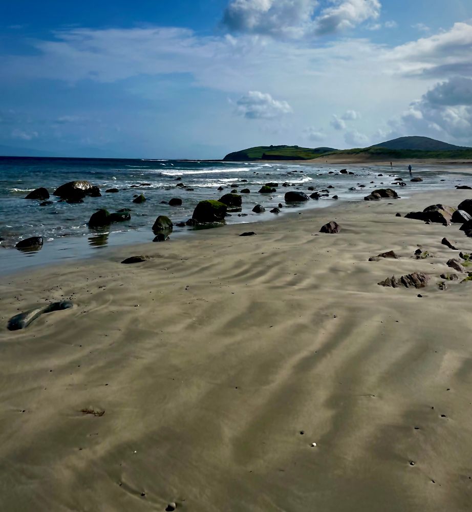 Carrowmore Beach. The tide is out and the rocks that line the shoreline are showing. The sky is mostly bright blue with some   White wispy cloud. In the background are low lying mountains. 