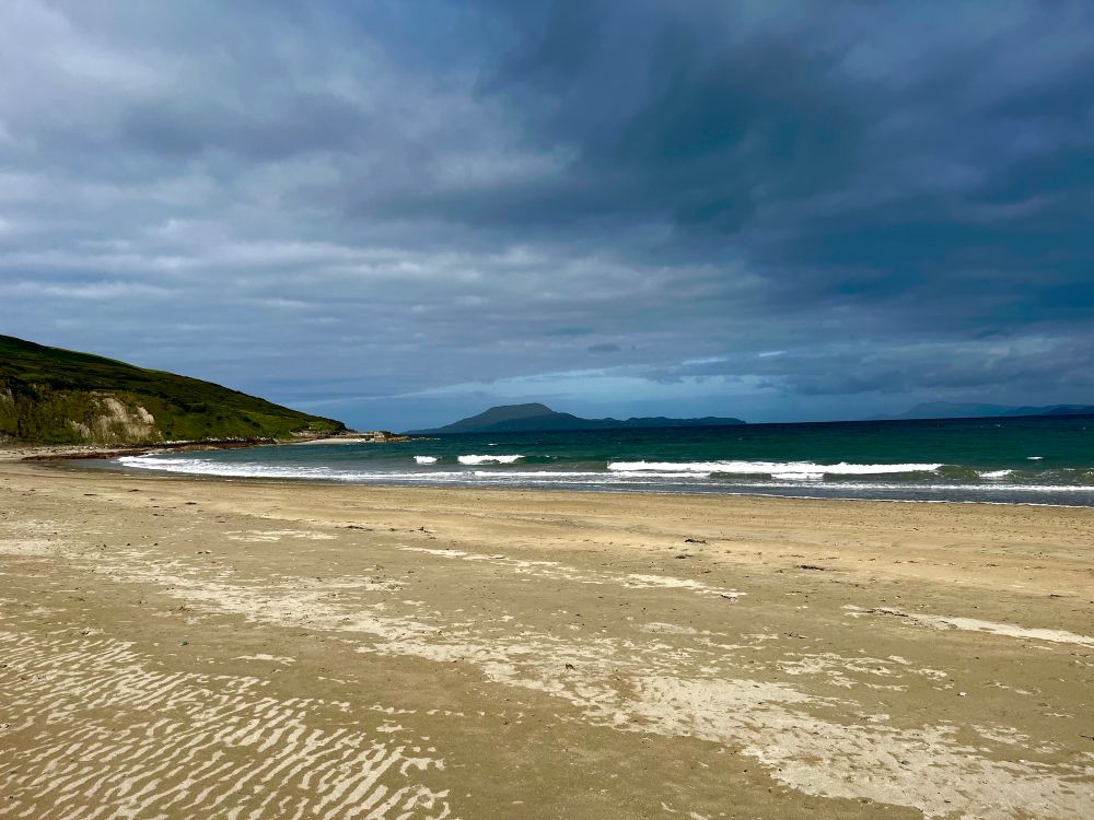 A view from the beach across golden sand and dark looking waters over to Clare Island in the distance. It is dark and murky looking. The sky gives the sense of the dark storm clouds rolling in as they cover have the sky in the image. 