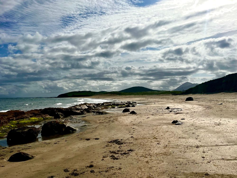A panoramic view of a beach in Mayo. The picture is framed with sand to the right and the touch of the shore line to the left incorporating the rocks and which is spattered with green. The clouds are mottled and textured 