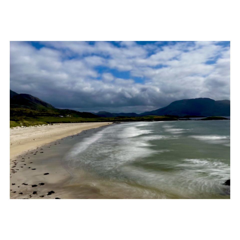 A scenic photograph of a beach with the water to the right and golden sand to the left. The sky is mottled with cloud and with blue sky poking through. In the background are mountains in various shades of green 