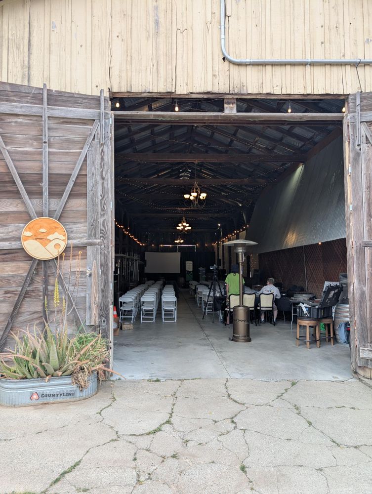 The front outside wall of a barn, covered in cream-coloured paint. Inside the barn, you can see a space set for a conference, with rows of chairs in the midfield and a projector screen. Two people are sitting at a desk. Hanging on the door (propped open) is a wooden North Bay Python logo. 