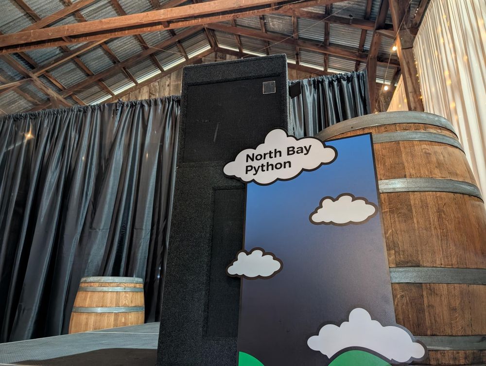 A photo of the lectern front featuring the green hills, blue skies, and fluffy white clouds of the North Bay Python branding leaning against a wine barrel on stage.
