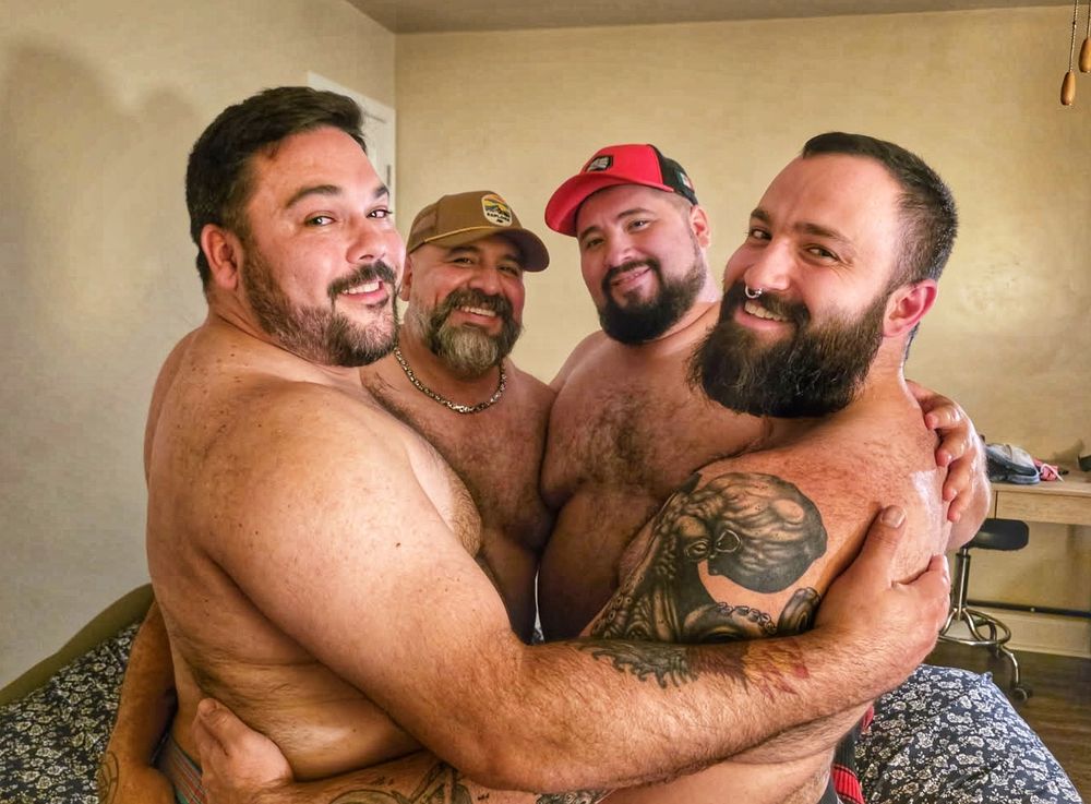 Four shirtless beautiful, bearded, beary men, all embracing and smiling at the camera.