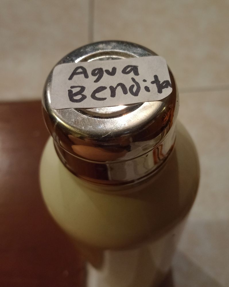 An off white, metallic water bottle with a silver lid and a sticker with "agua bendita" (spanish for holy water) written with sharpie.