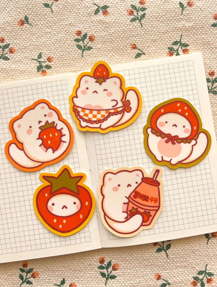 Cute cat-themed stickers with strawberry and milk designs are laid out on a graph paper notebook. The background features a fabric with small floral patterns.